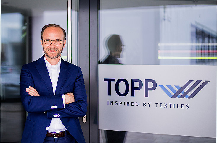 Geschäftsführer Dr. Stefan Topp im Anzug mit verschränkten Armen vor dem Haupteingang mit groß sichtbarem Unternehmenslogo.